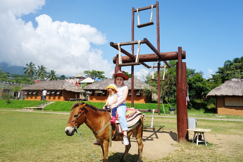 Girl riding a horse with traditional village huts and trees in the background at Santichon Village.