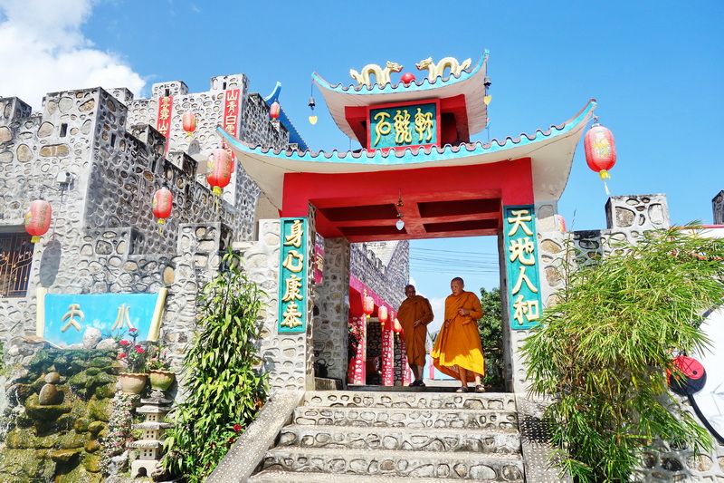 Two monks standing at the entrance of a temple with stone walls, lanterns, and a blue sky at Mae Hong Son.