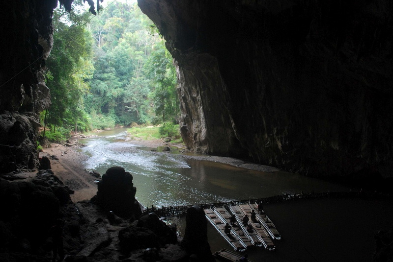 View from inside a dark cave with a river and green trees outside at Tham Lod Cave.