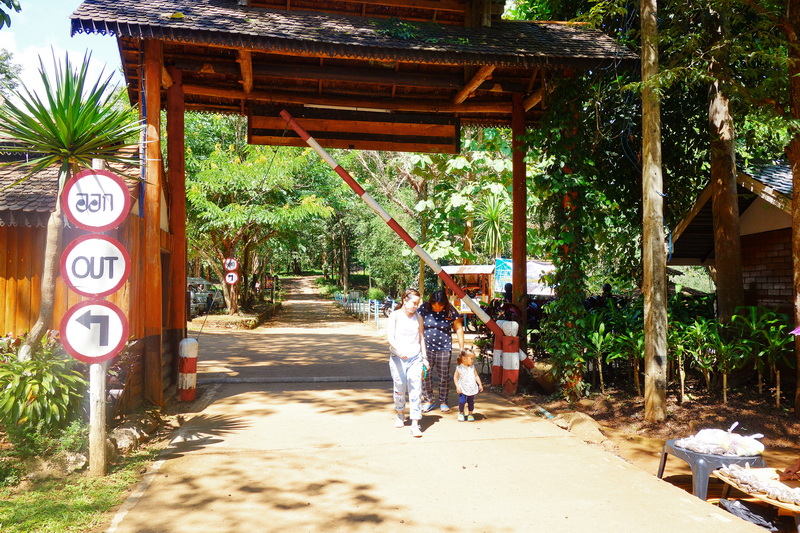 People walking under a wooden gate with barrier arm amid greenery and sunlight at Tham Lod Cave.