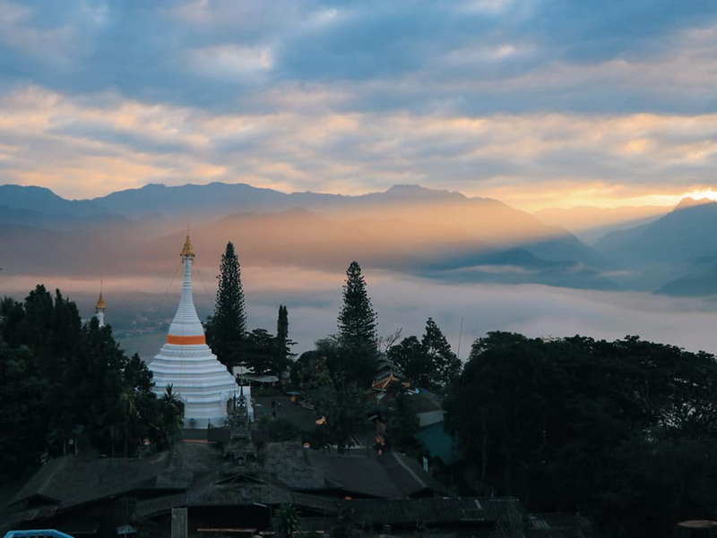 White stupa with gold accents among trees and mountains under cloudy sky at Wat Phra That Doi Kong Mu.