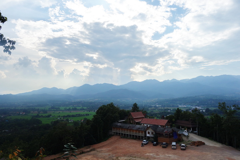 Scenic viewpoint with mountains, fields, building, and parked cars under a cloudy sky at Wat Phra That Mae Yen.