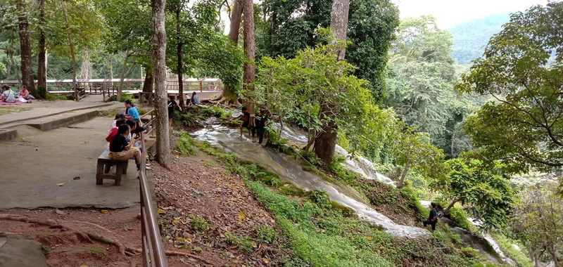 People relaxing and walking in a shaded forest area with a small stream at Buatong Waterfall.