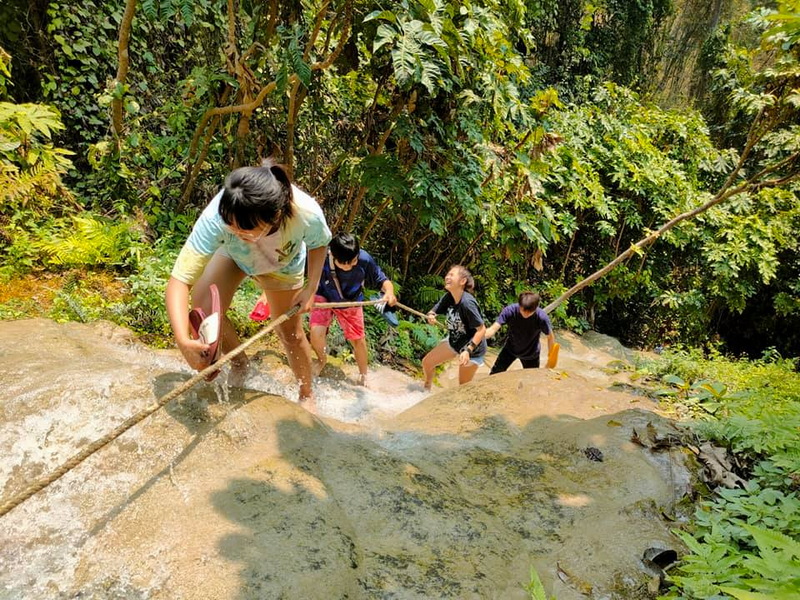 Children Climbing Wet Rock at Buatong Waterfall Children climbing a wet rocky terrain with a safety rope in a dense forest at Buatong Waterfall.