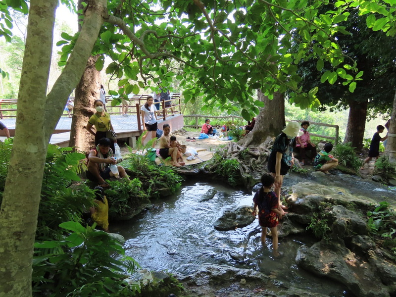People enjoying relaxing by a small river surrounded by trees and rocks at Buatong Waterfall.