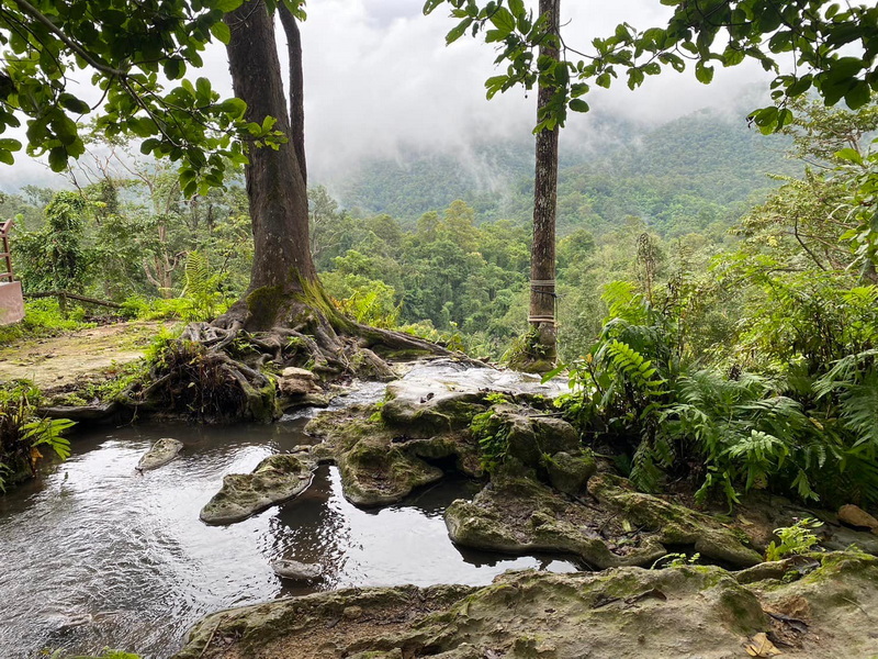 Small stream flowing over rocks with lush greenery and misty mountains in background at Buatong Waterfall.