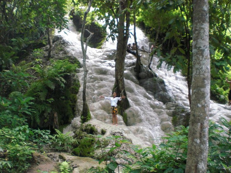 Person with arms outstretched at a cascading waterfall amidst dense jungle greenery at Buatong Waterfall.