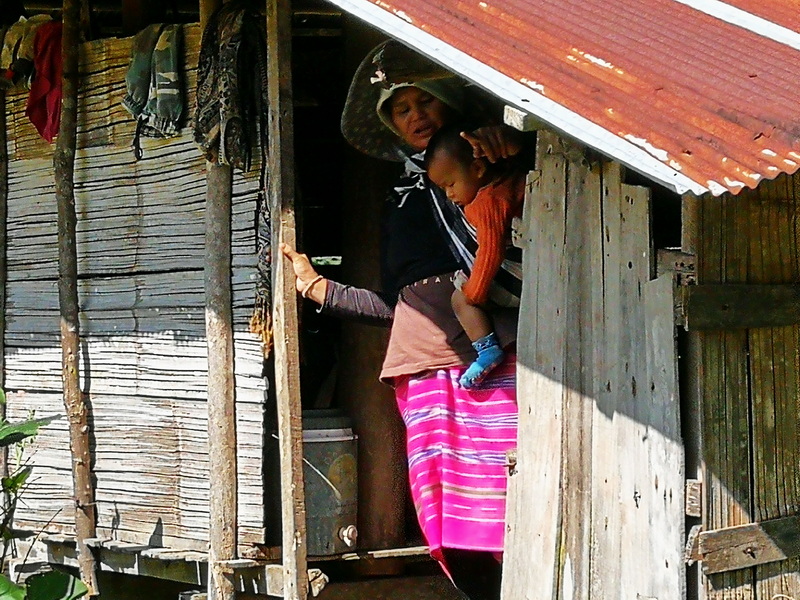 Woman holding a child outside a wooden hut with a metal roof at Ban Mae Klang Luang.