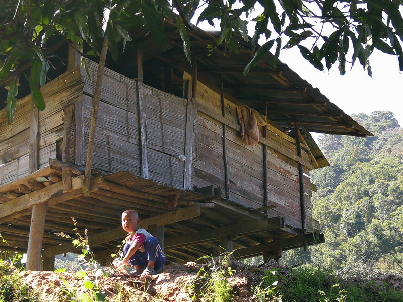 Young boy sitting outdoors in front of a wooden stilt house amid trees and greenery at Mae Klang Luang Village.