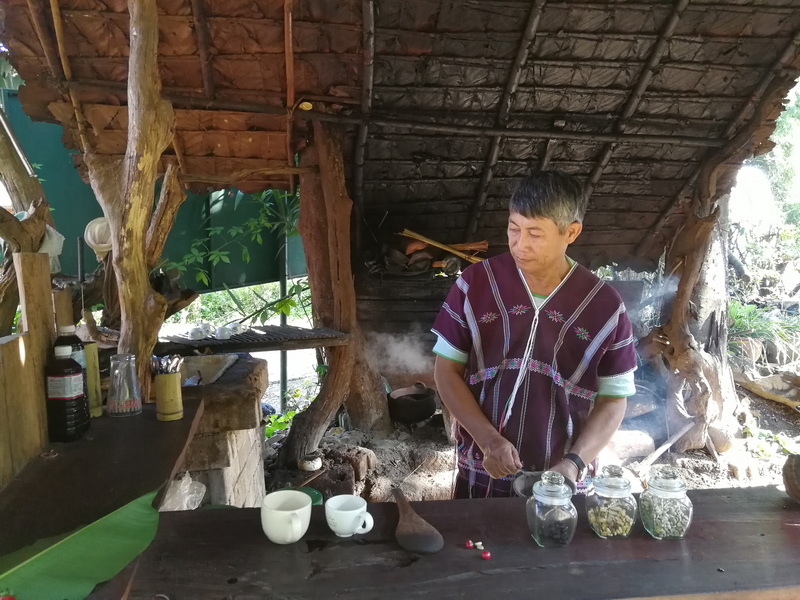 Traditional Wooden Kitchen Scene at Ban Mae Klang Luang Person in traditional attire in a rustic wooden kitchen with jars and cups inside a nature setting at Ban Mae Klang Luang.