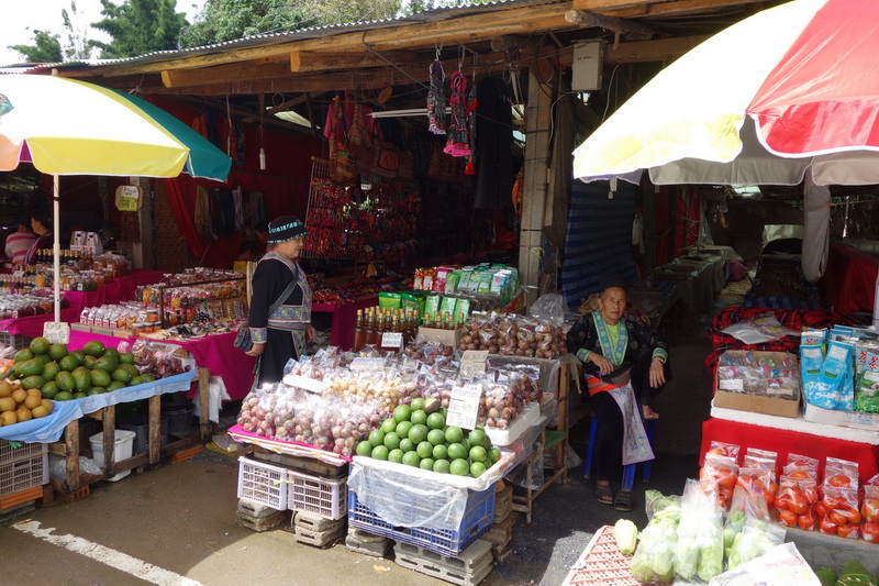 Market stall with fruits and goods under umbrellas with two women in traditional attire at Doi Inthanon National Park.