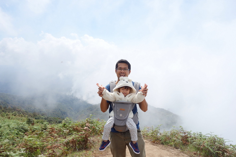Man with a child in a carrier on a mountain trail with clouds and greenery at Kew Mae Pan Nature Trail.