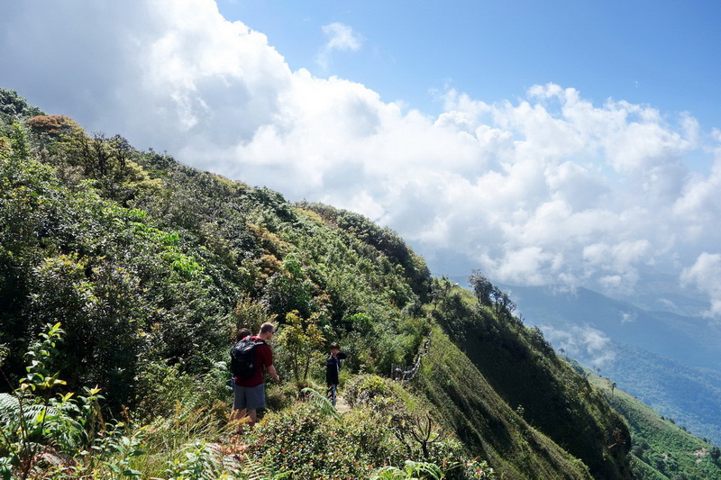 Hikers walking on a mountain trail with lush greenery and cloudy sky above at Kew Mae Pan.