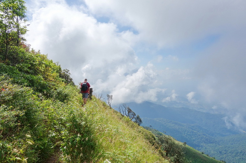 Hiker with backpack walking on a grassy trail with mountain view and clouds behind at Doi Inthanon National Park.