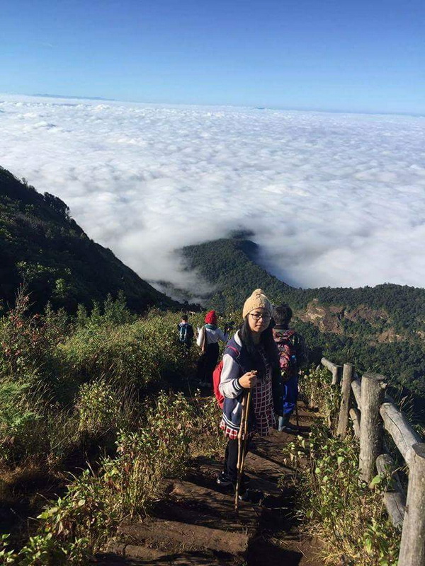Hikers walking along a mountain trail above clouds with forested peaks in the distance at Kew Mae Pan.