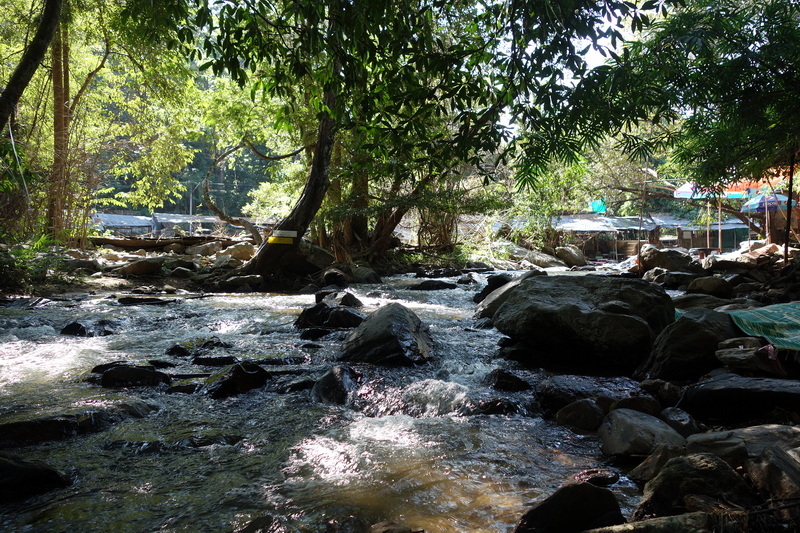 A rocky stream in a lush forest with sunlight shining through trees at Mae Klang Waterfall.