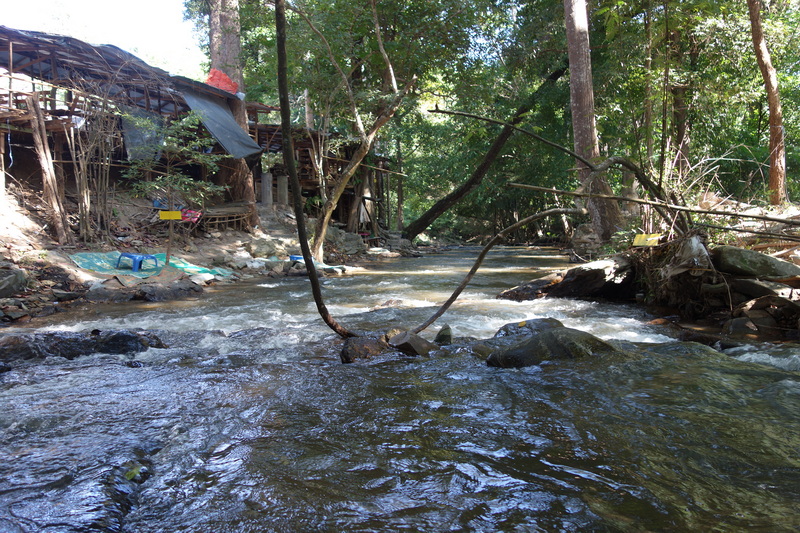 A river flowing through a forest with wooden structures on the bank at Mae Klang Waterfall.