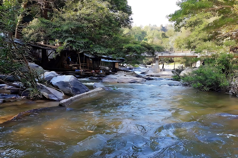 River flowing with rocks and trees, bridge in background, near Mae Ya Waterfall at Mae Klang Waterfall.