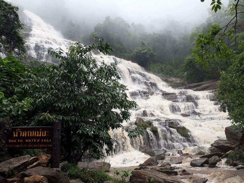 A multi-tiered waterfall flowing over rocks with lush green trees and mist in the background at Mae Ya Waterfall.