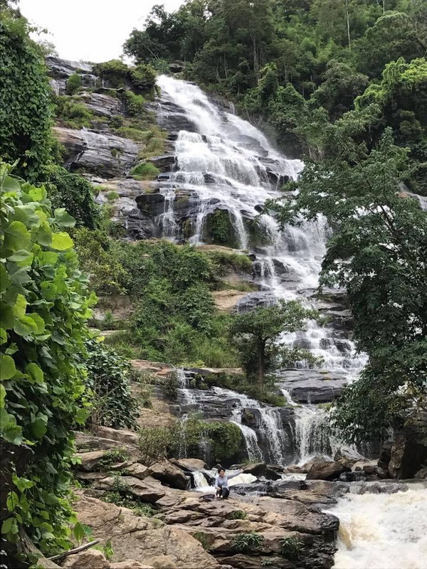 Waterfall flowing over rocks surrounded by green foliage with a person sitting nearby at Mae Ya Waterfall.