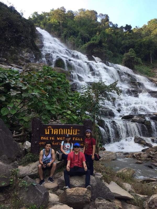 Hikers at Mae Ya Waterfall Four people stand near a sign at Mae Ya Waterfall with a cascading waterfall behind them.