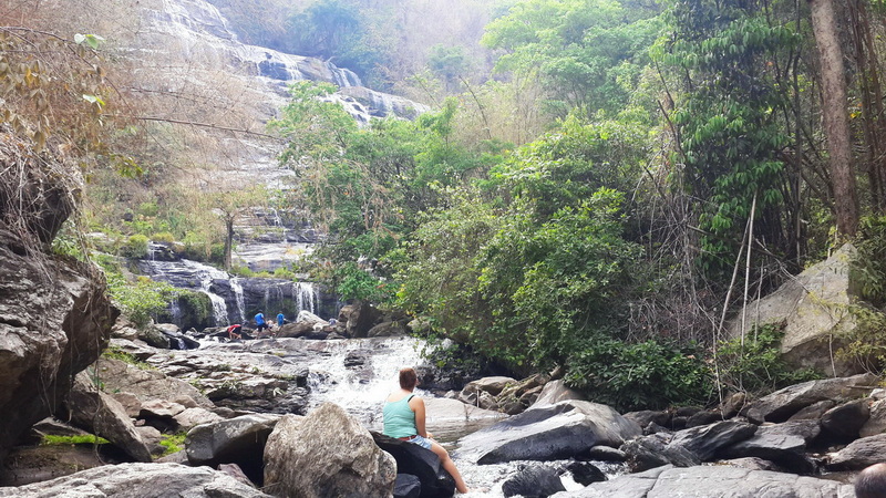 Serene Streamside Scene at Mae Ya Waterfall Woman sitting on rocks beside flowing stream with greenery around at Mae Ya Waterfall.
