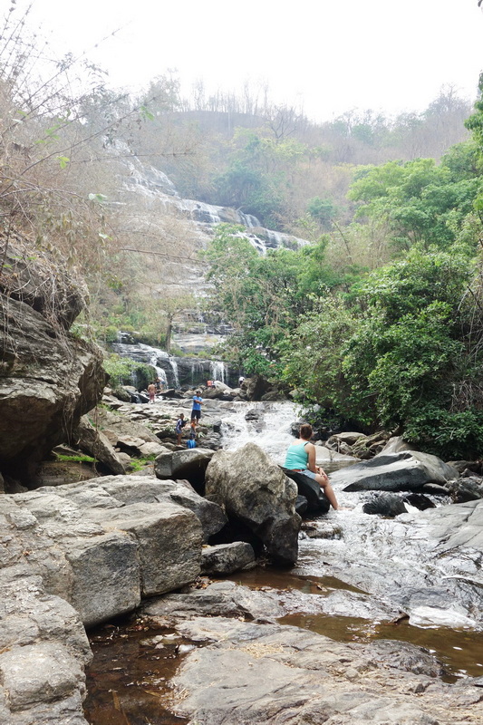 People sitting on rocks and playing in a shallow river with a waterfall and green trees behind at Mae Ya Waterfall.