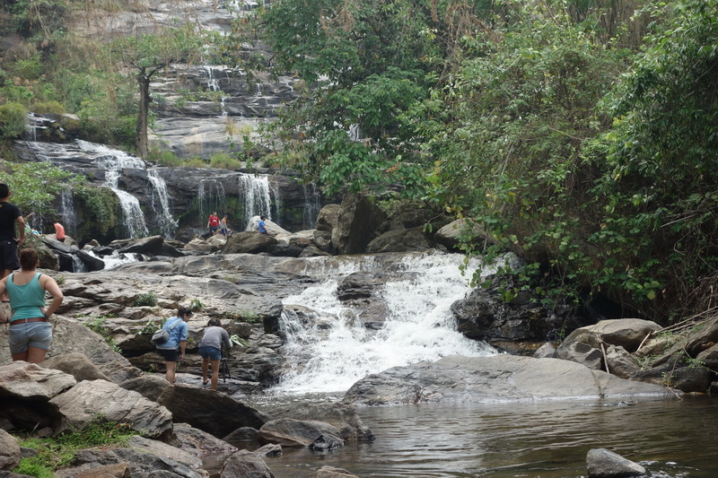 Visitors walking on rocks near a multi-tiered waterfall with lush trees around at Doi Inthanon National Park.