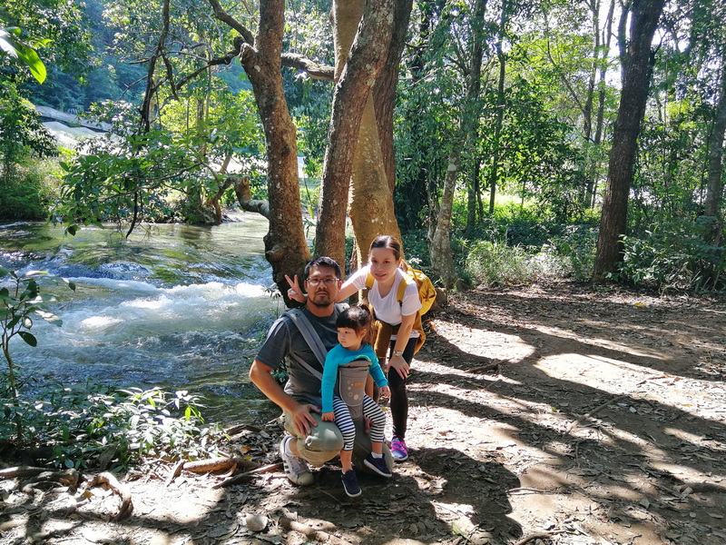 Family posing on a sunlit forest trail beside a flowing river and trees at Pha Dok Siew Nature Trail.