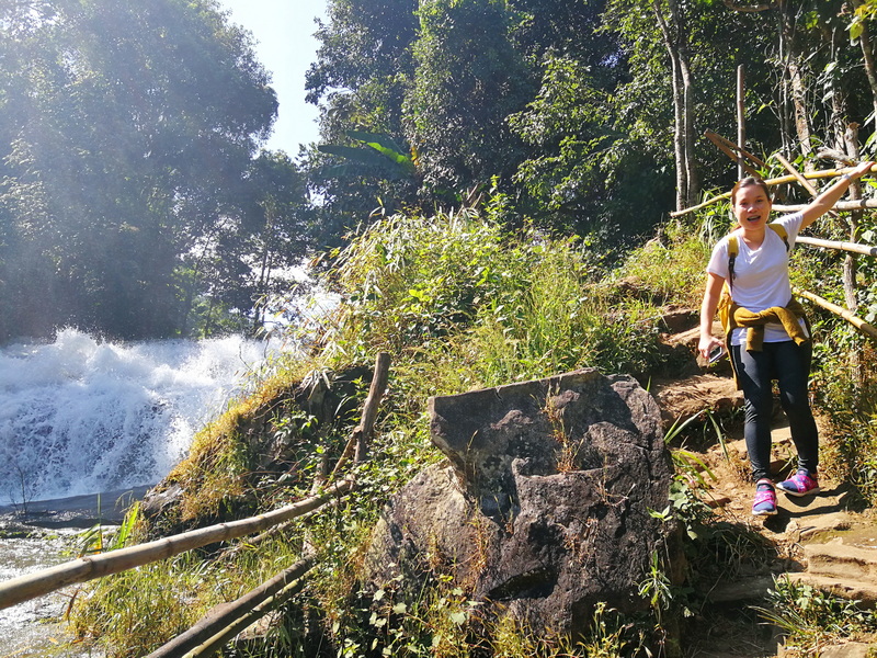 Woman smiling while hiking on a rocky trail near a waterfall in a lush forest at Pha Dok Siew Nature Trail.