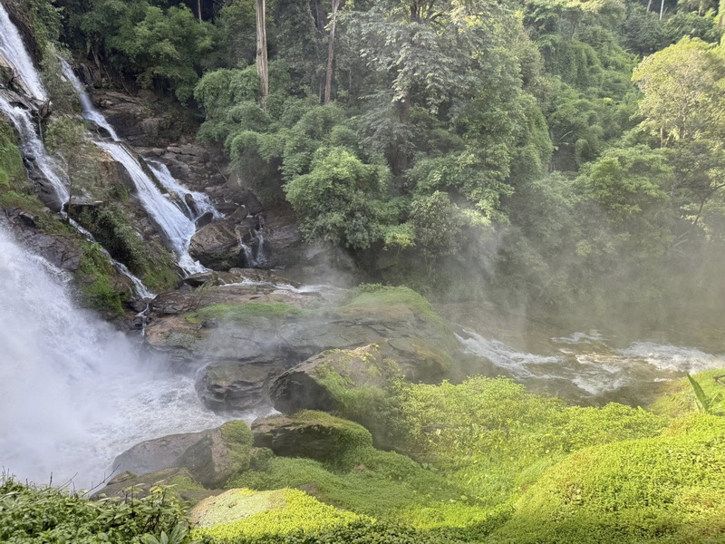 Water cascading over rocks amidst dense green trees and misty surroundings at Wachirathan Waterfall.