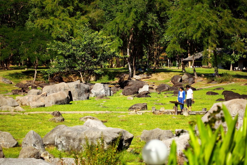 Two people standing near rocks and grass among trees in a scenic park at Fang Hot Springs.