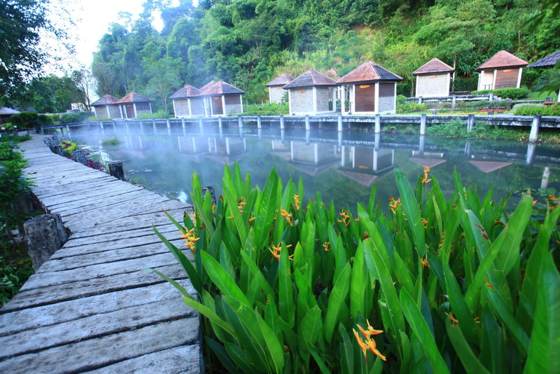 Wooden walkway beside steaming hot spring pond with small pavilions and green hills at Fang Hot Spring.