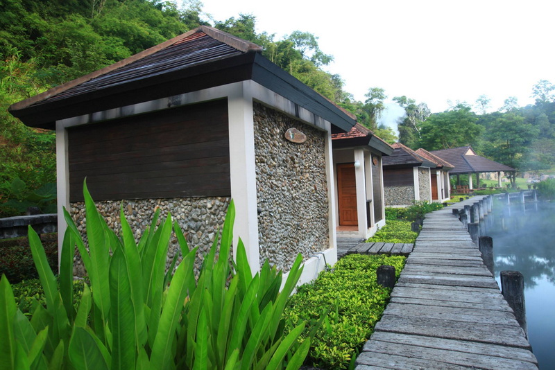 Small buildings with stone and wood walls beside a wooden path over water, lush trees in background at Fang Hot Springs.