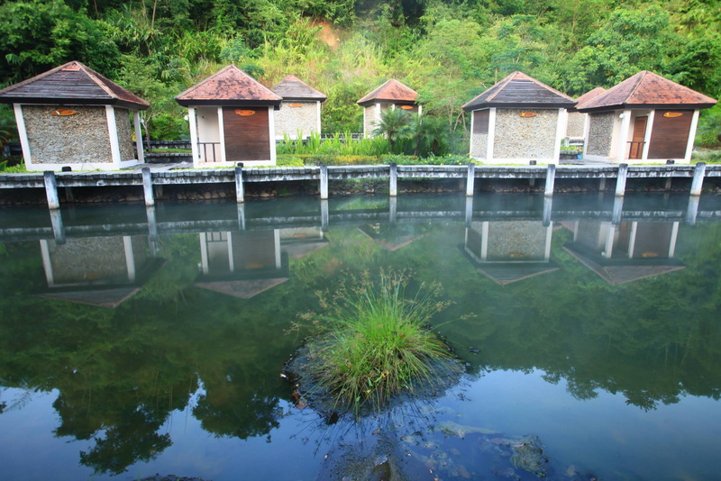 Six small buildings along a canal with greenery and reflections in the water at Fang Hot Springs.