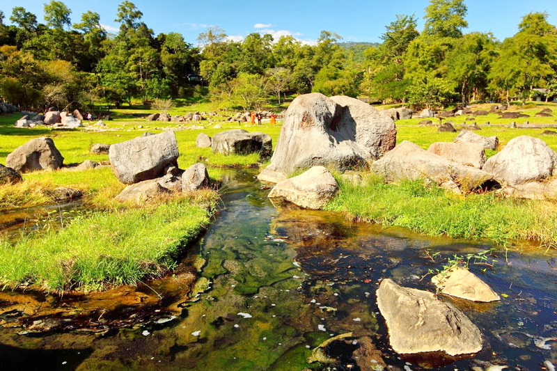 A stream winding through a grassy field with large rocks and trees in the background at Fang Hot Spring.