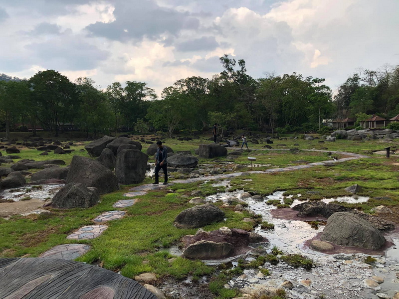 People walking on a stone pathway surrounded by grass, rocks, and trees at Fang Hot Springs.