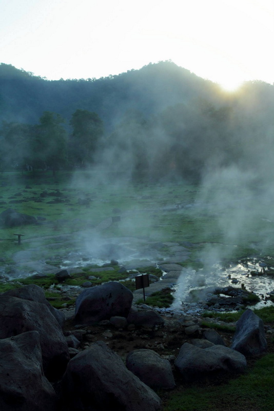 Steam rises from hot spring rocks with trees and mountains in the background at Fang Hot Spring.