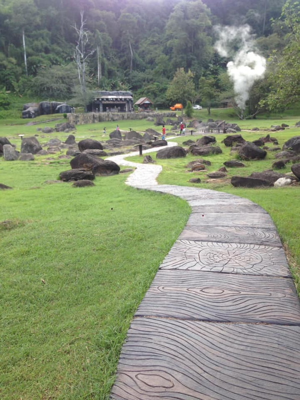 Curved stone pathway through a green grassy field with rocks and trees in the background at Fang Hot Spring.