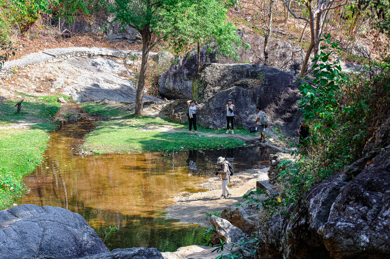People standing beside a small river with trees and rocks in a natural landscape at Huay Kaew Waterfall.