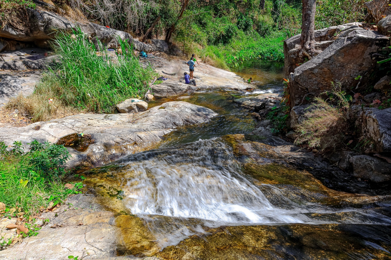 Streamside Relaxation in Nature at Huai Kaew Waterfall People relaxing on rocks beside a flowing stream surrounded by trees and plants at Huai Kaew Waterfall.