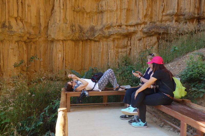 Three women sitting on benches beside a rock cliff, two using phones, one lying down at Phar Choi.