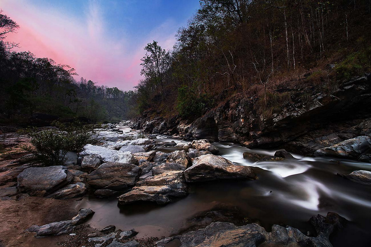 River flowing over rocks in a forested area with colorful sky at National Parks in Chiang Mai.