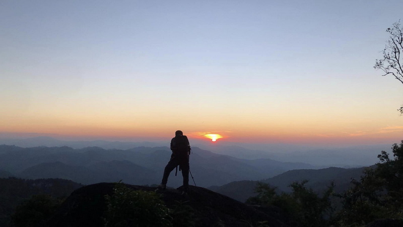 Sunset View from Mountain Peak at Pha Daeng National Park Person standing on a rock ledge capturing a sunset over layered mountain ranges at Pha Daeng National Park.