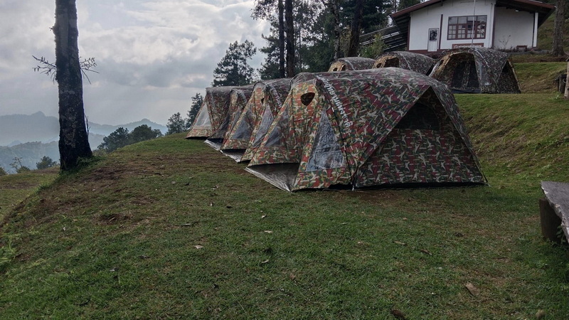 Camping Scene in Nature at Pha Daeng National Park Camouflage tents set up on a grassy hillside with trees and a house nearby at Pha Daeng National Park.