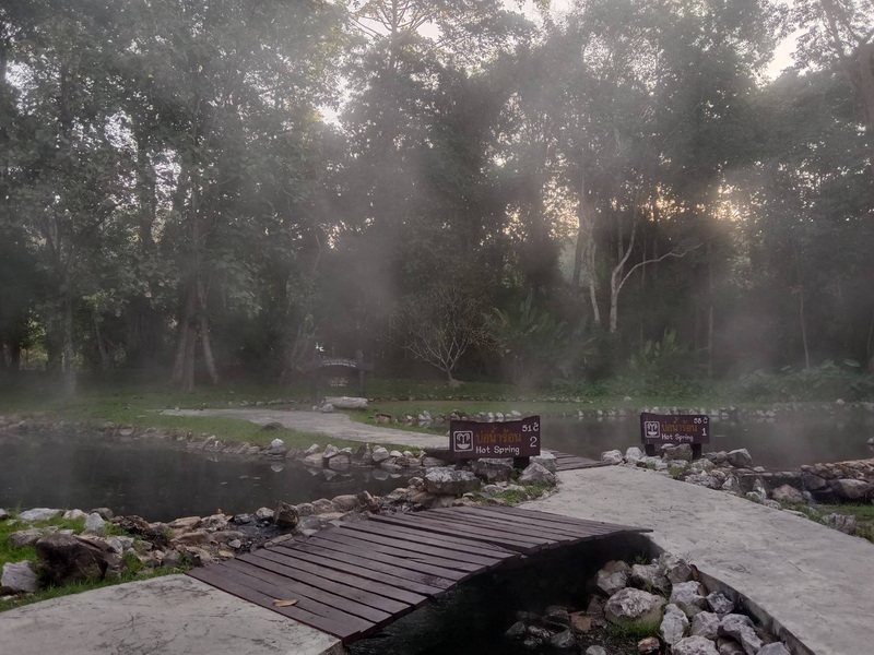 Steam rises from hot springs with trees and rocks around, signs in view at Pha Daeng National Park.