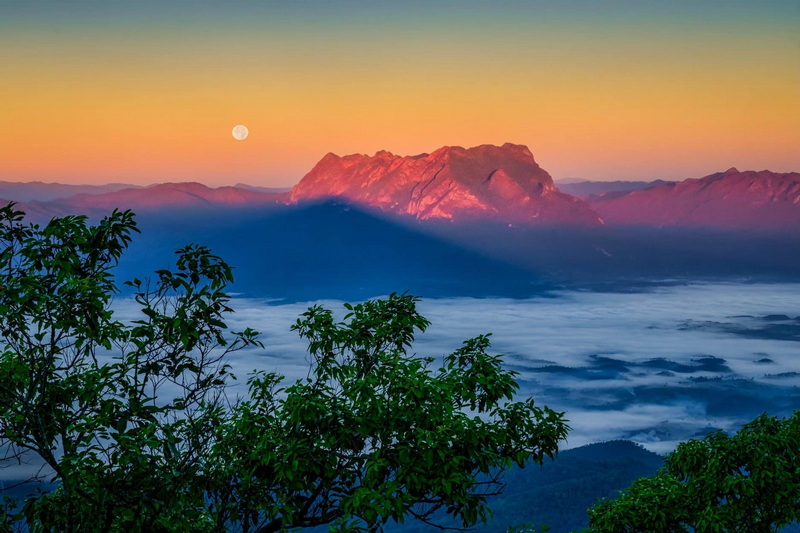 Sunlit mountain range with full moon above clouds and green foliage in foreground at Sri Lanna National Park.