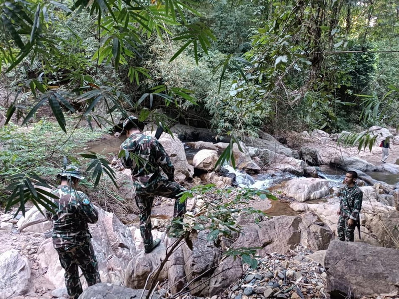Three individuals in camouflage uniforms explore a rocky stream in a dense forest at Si Lanna National Park.