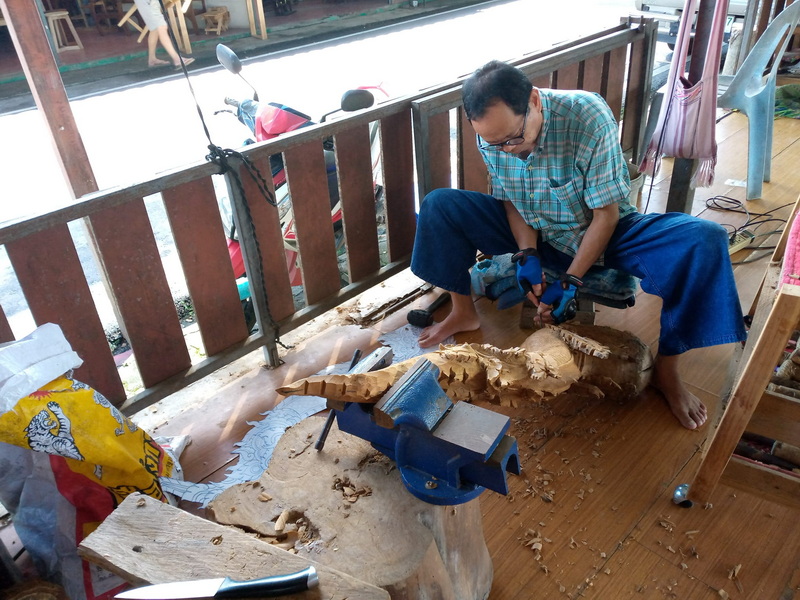 Wood Carving Workshop Scene at Baan Tawai Man carving wood with a power tool on a wooden workbench in a workshop setting at Baan Tawai.
