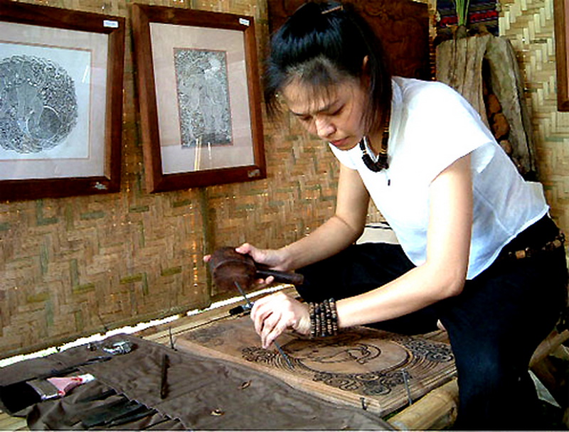 Wood Carving Inside Rustic Workshop at Baan Tawai Woman carving wood on a worktable inside a rustic room with framed art on the wall at Baan Tawai.